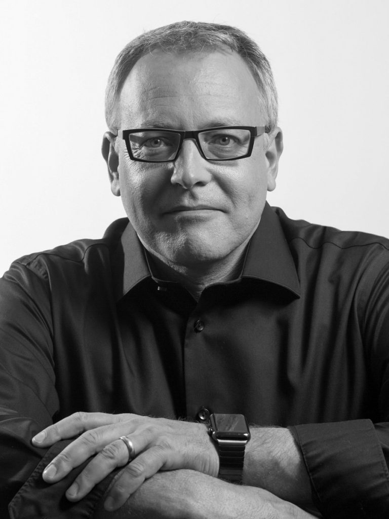 A black and white headshot of a man wearing glasses, seated