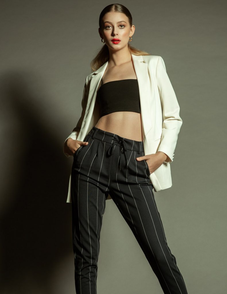 A fashion model posing in a white jacket, black crop top, and dark pinstripe panst