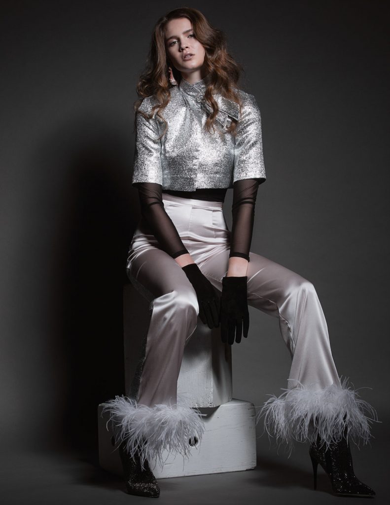 A fashion model posing seated in silver jacket and pants with feather accents